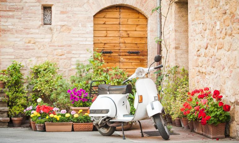 Scooter blanc garé devant une porte avec des plantes et fleurs colorées.